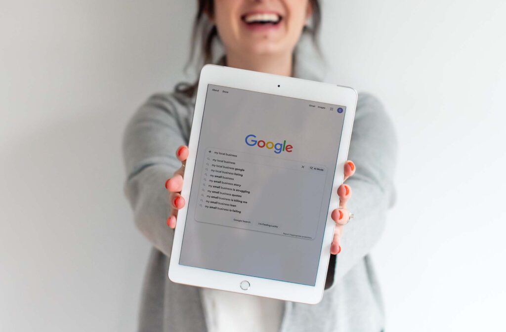 Woman smiling while holding an iPad showing Google search results for "my local business" with AI Mode visible